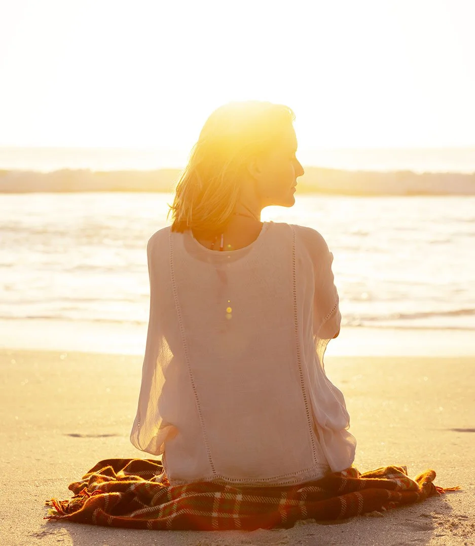 Woman sitting on a beach at sunset, facing the ocean, with her hair illuminated by the sunlight, seated on a blanket.