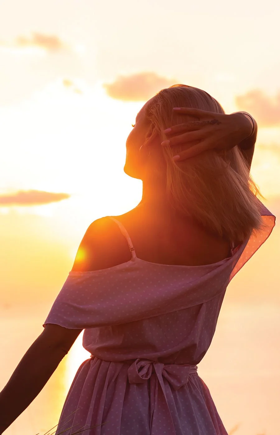 A woman with blonde hair wearing a pink off-shoulder dress, standing outdoors during sunset, with her hand in her hair.