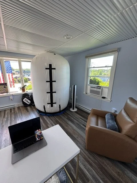 A large white coffee bean-shaped object with black markings stands in a living room next to a window air conditioning unit. The room has wood flooring, a beige armchair, a white table with an open laptop, and a window with an American flag decoration in the background.