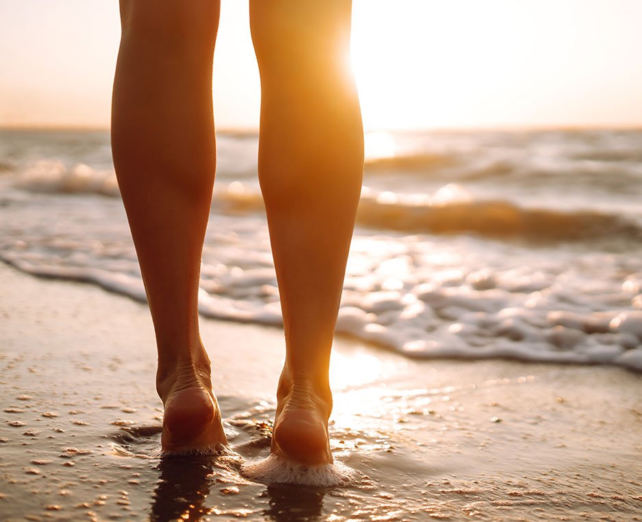 Close-up of a person's legs and feet walking on the wet sand at the beach with ocean waves in the background during sunset.