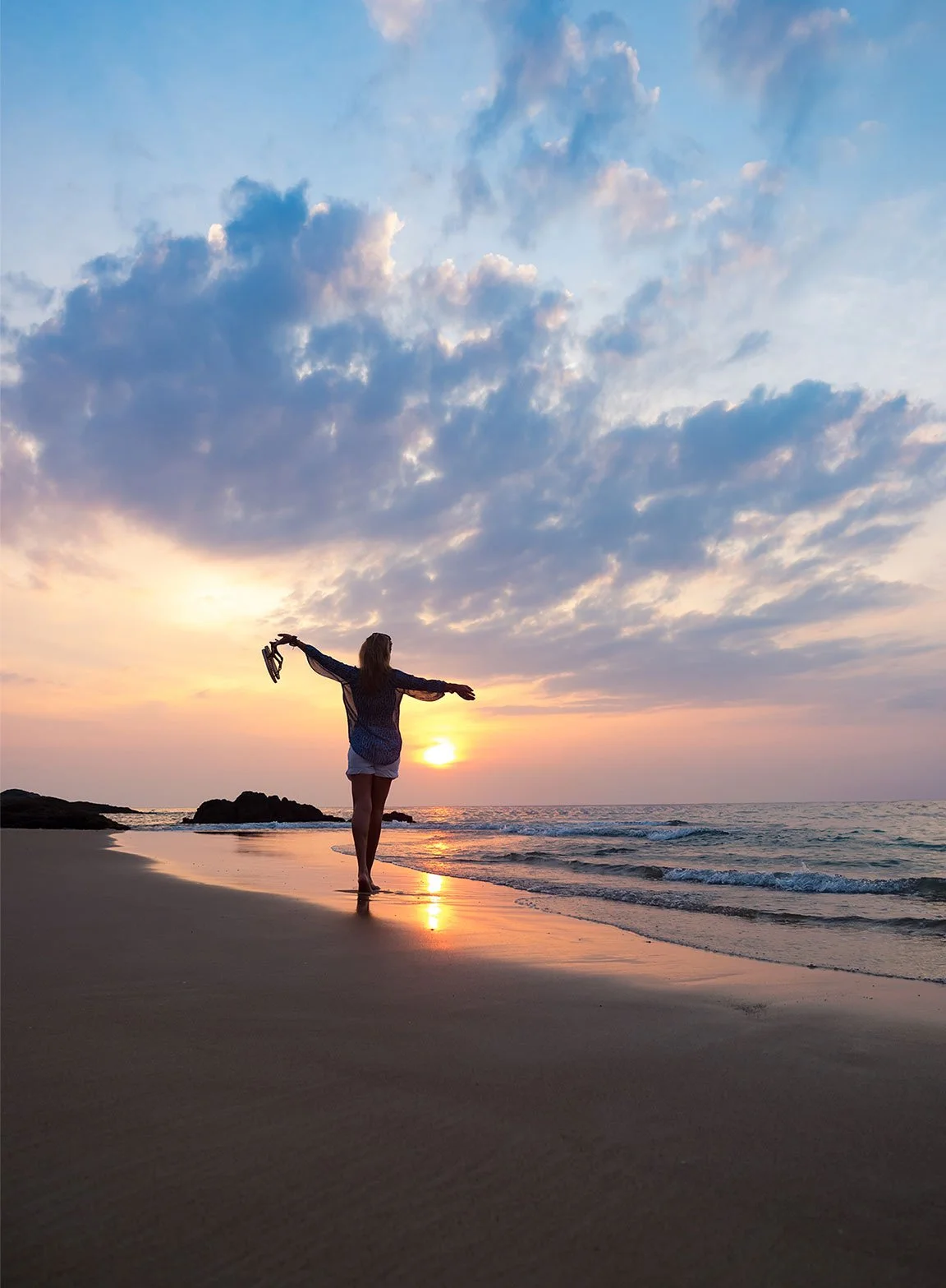 A woman standing on a beach at sunset, facing the ocean with arms stretched out, holding shoes in one hand, with a colorful sky and clouds overhead.