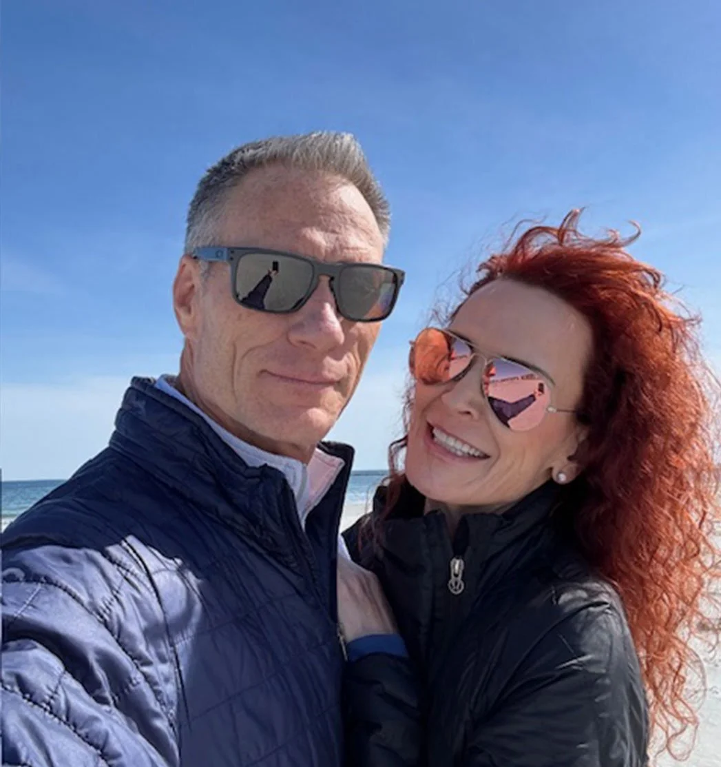 A smiling couple taking a selfie on the beach, both wearing sunglasses and jackets, with the ocean and a clear blue sky in the background.