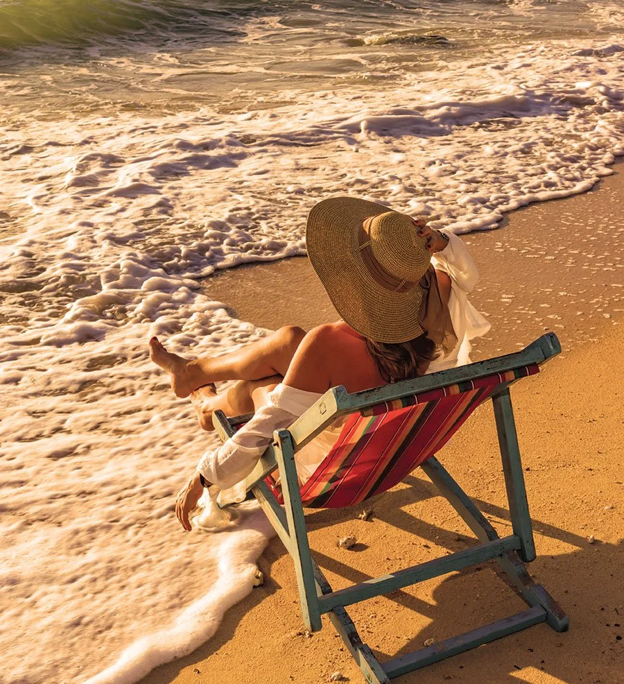 A woman wearing a large sunhat and white clothing relaxing on a striped beach chair by the ocean at sunset, with waves lapping at the shore.