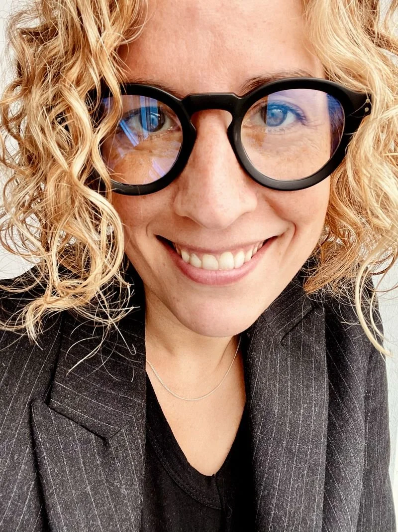 A woman with curly blonde hair wearing large round black glasses smiles at the camera, dressed in a dark pinstripe blazer and black top.