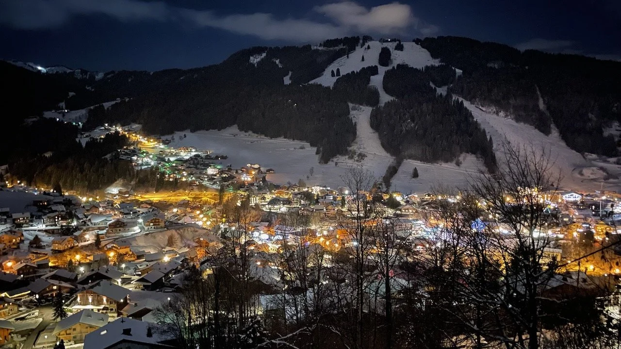 Nighttime view of a snow-covered mountain village illuminated with streetlights and house lights, with ski slopes and dark forested mountains in the background.