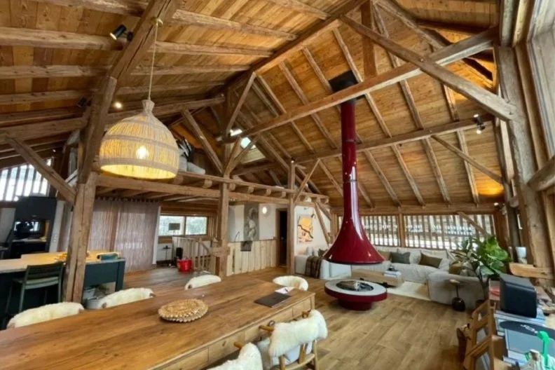 Spacious living area with exposed wooden beams, a large red fireplace, and modern furniture. Pendant lighting, lights on high beams.