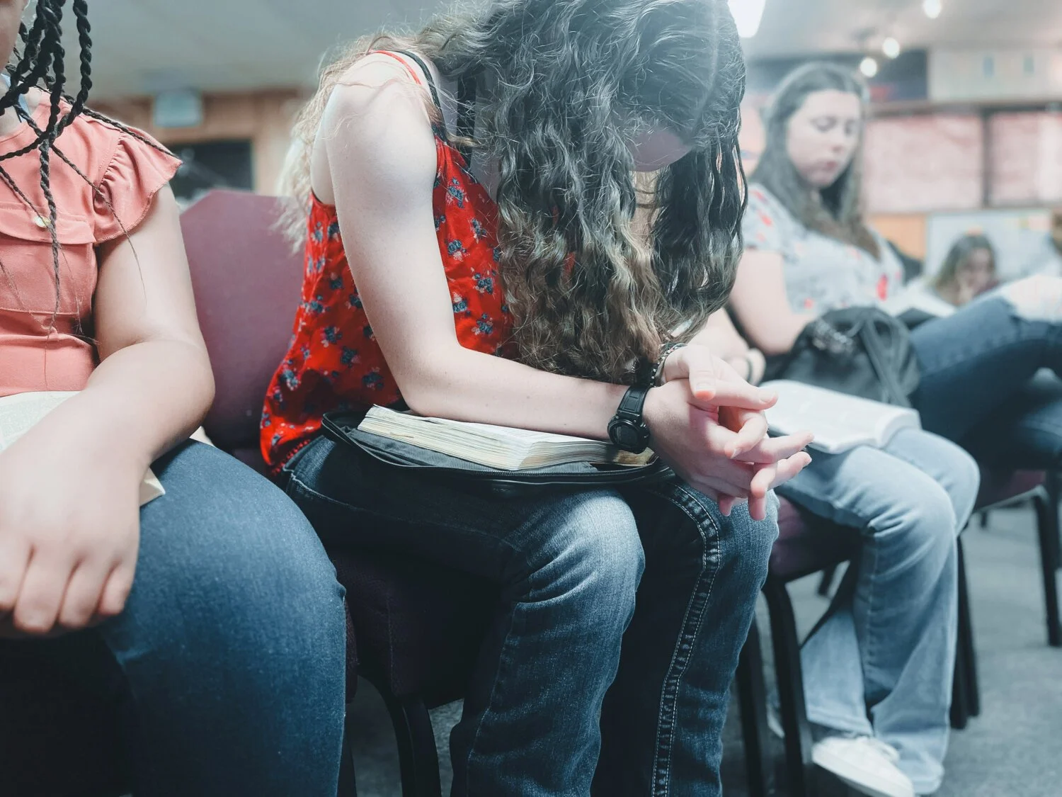People sitting in a row at a church or meeting, with one woman with long curly hair and a red floral top with hands clasped, sitting with a Bible on her lap, in a prayer or meditative pose.