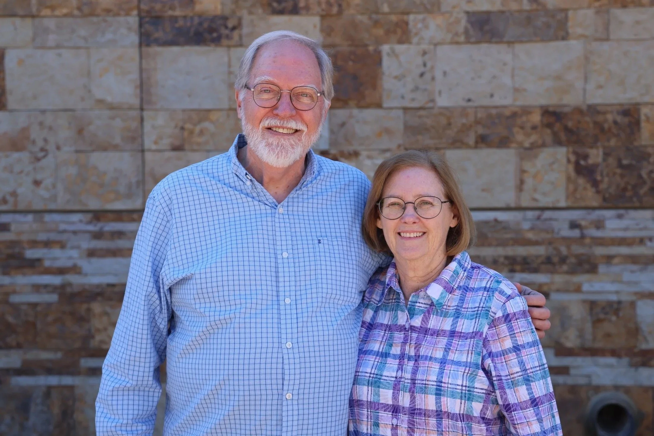 An elderly couple standing outdoors with a stone wall background, smiling, the woman wearing glasses and a colorful patterned top, the man with glasses and a black shirt.