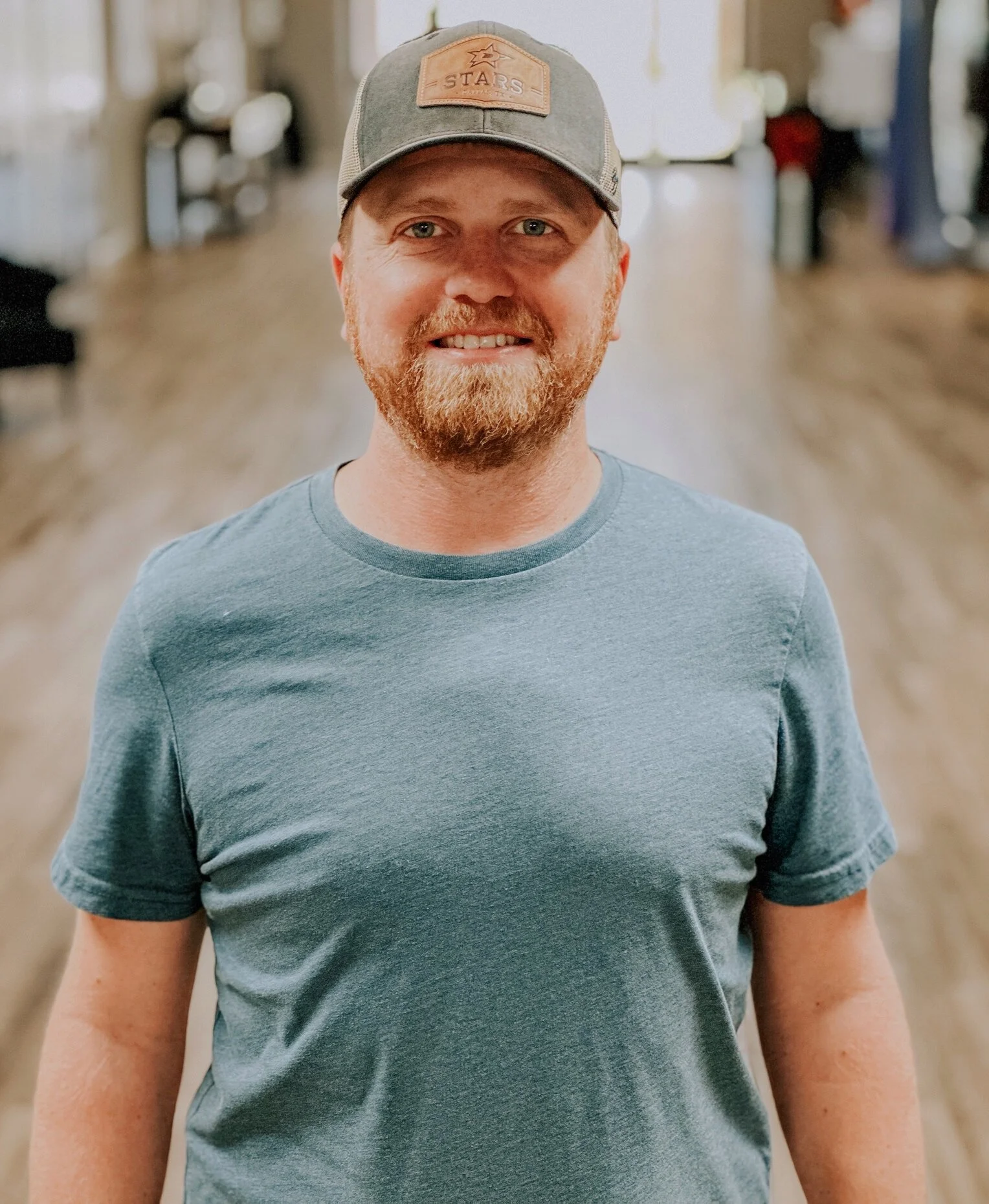 A person with fair skin, red beard and blue eyes wearing a gray t-shirt and a baseball cap, standing in a well-lit indoor space with blurred background.
