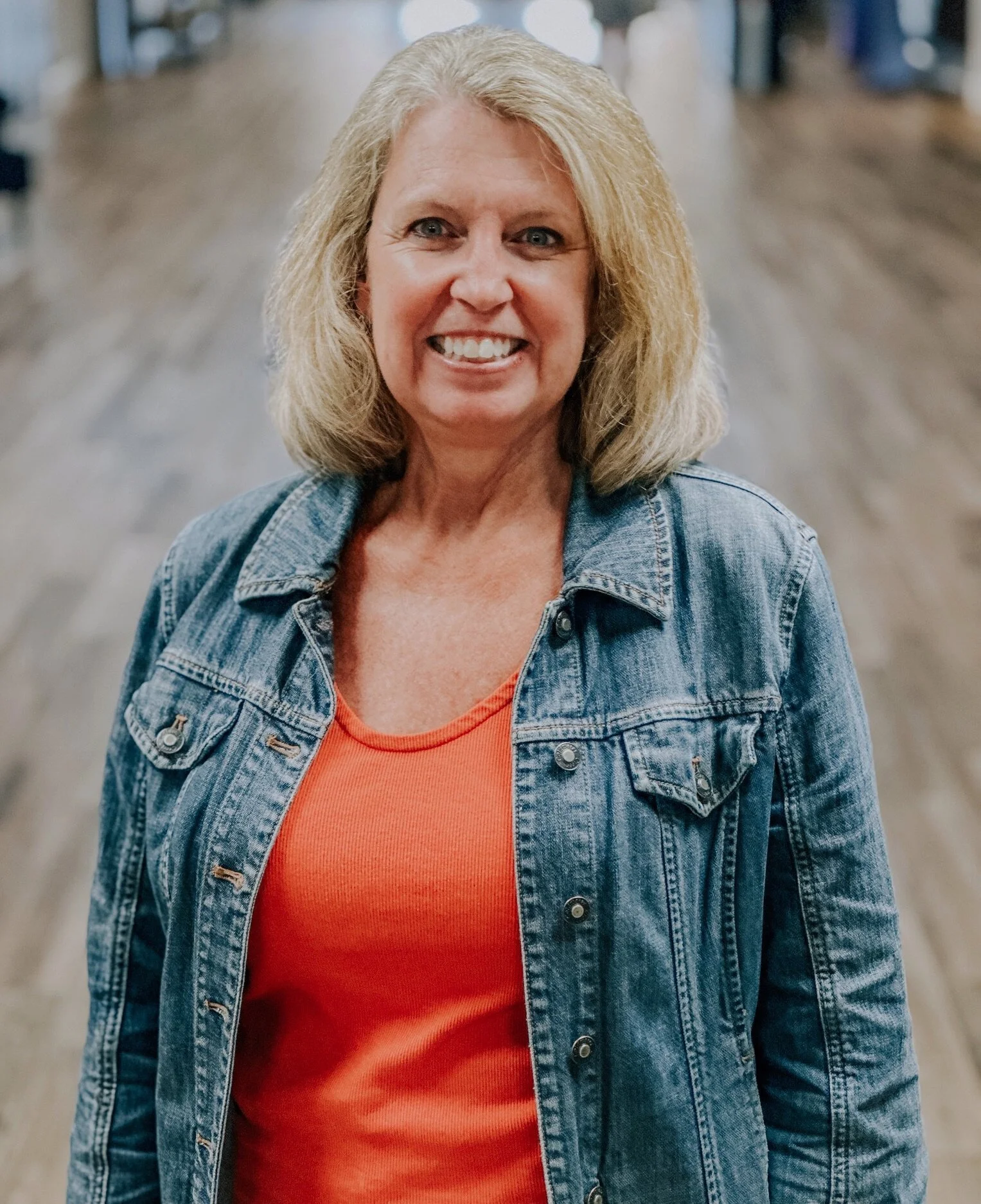 A middle-aged woman with blonde hair smiling and wearing a denim jacket and orange shirt, standing indoors.