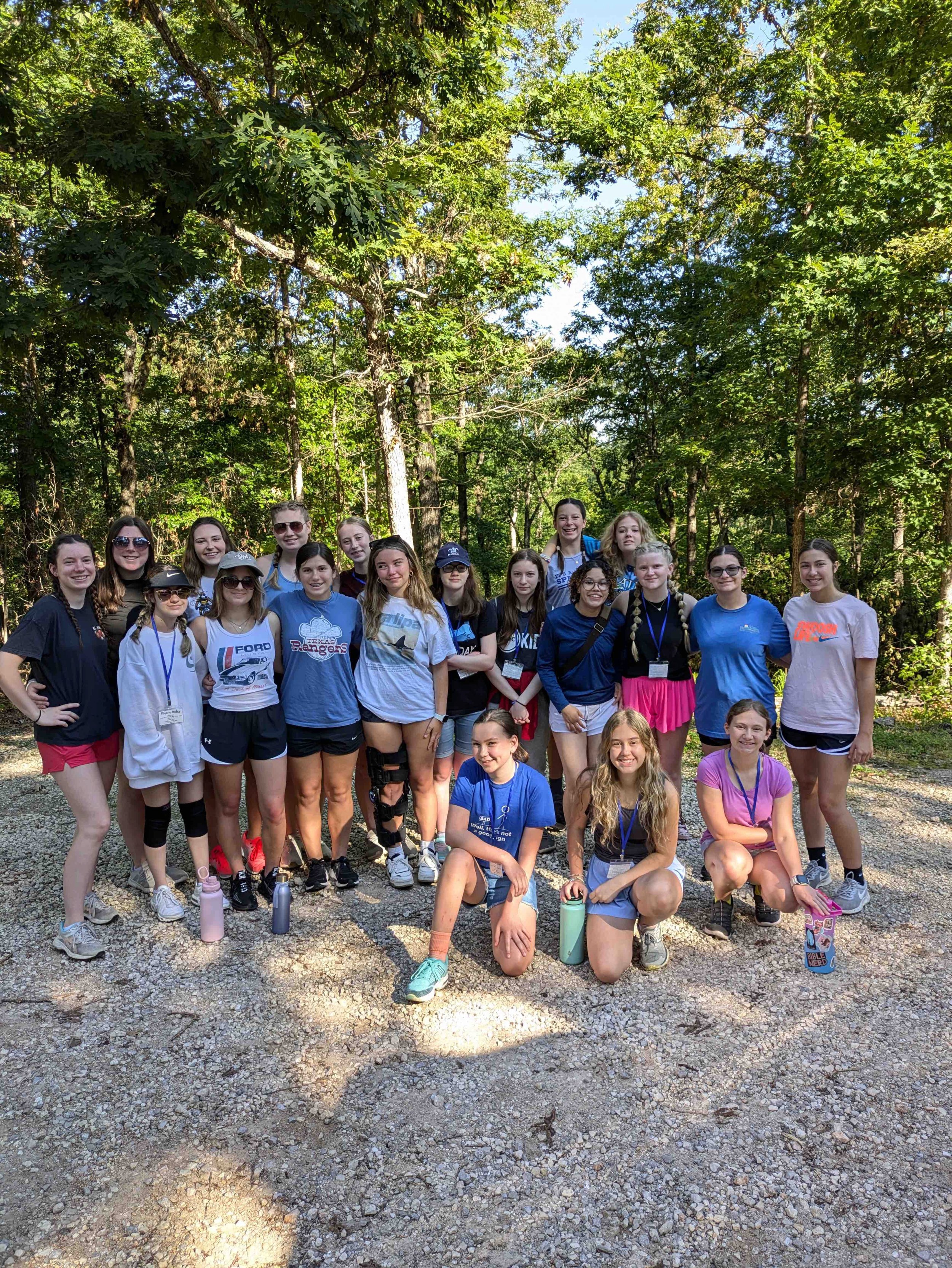 A group of young people, mostly girls, standing outdoors on a gravel path surrounded by trees. They are smiling and wearing casual athletic clothing, some with sunglasses and lanyards around their necks. The background features dense green foliage and trees in a forest or park setting.