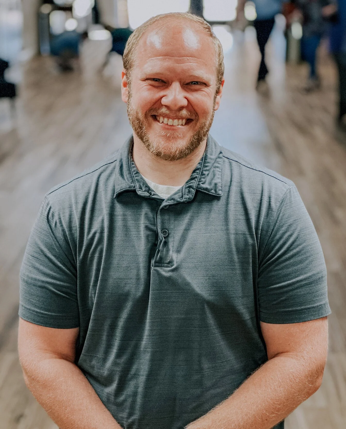 A smiling man with a beard and short hair, wearing a gray polo shirt, sitting in a well-lit indoor space with a blurred background of other people and wooden flooring.