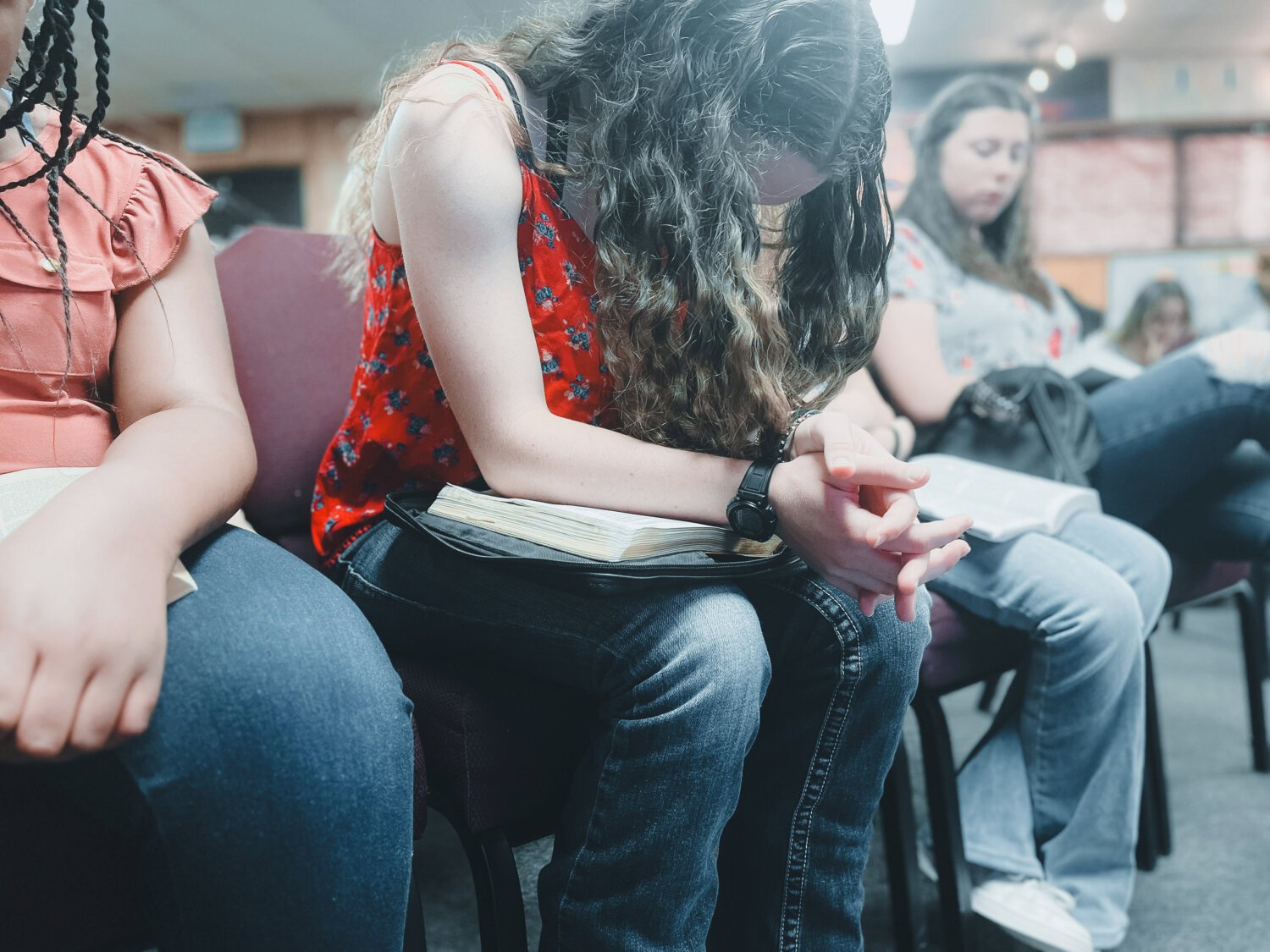 People seated in a room, engaged in prayer or reflection, with one woman having a prayer book on her lap, some with their eyes closed, in a worship setting.