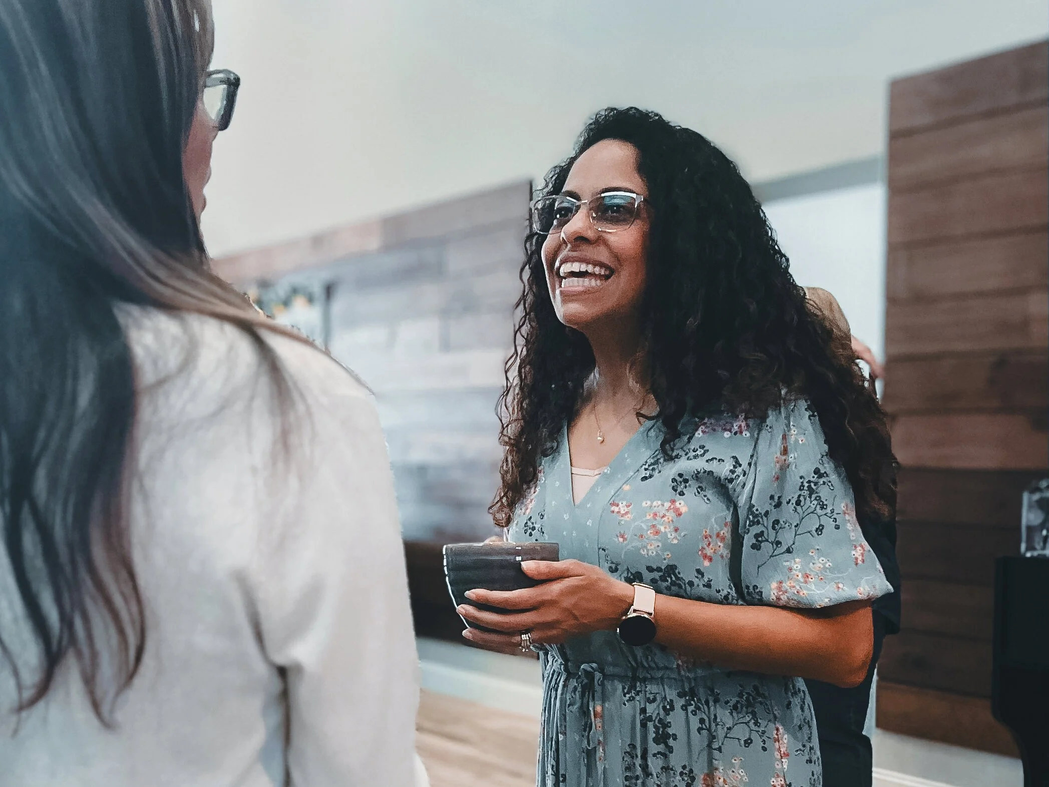 Two women engaged in conversation indoors, one with curly hair and sunglasses, holding a phone, and the other with straight hair and glasses.