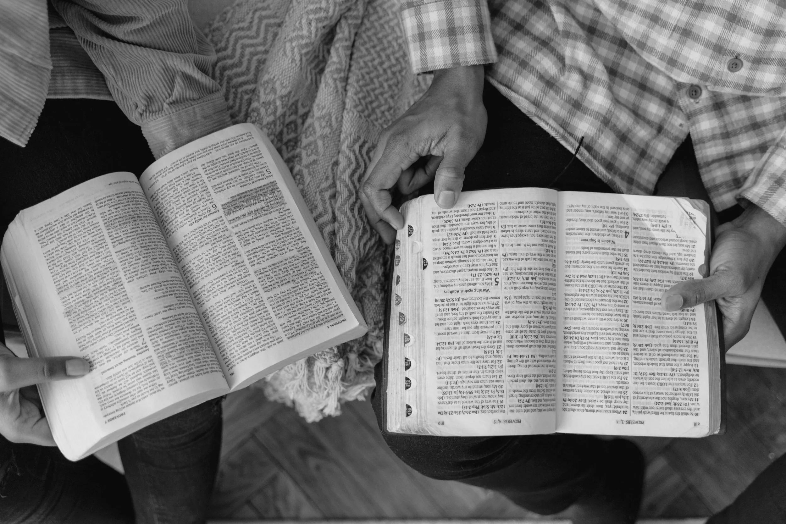 Two people sitting close together reading and sharing a book, with their hands near the open pages. The photo is in black and white, showing their casual clothing.