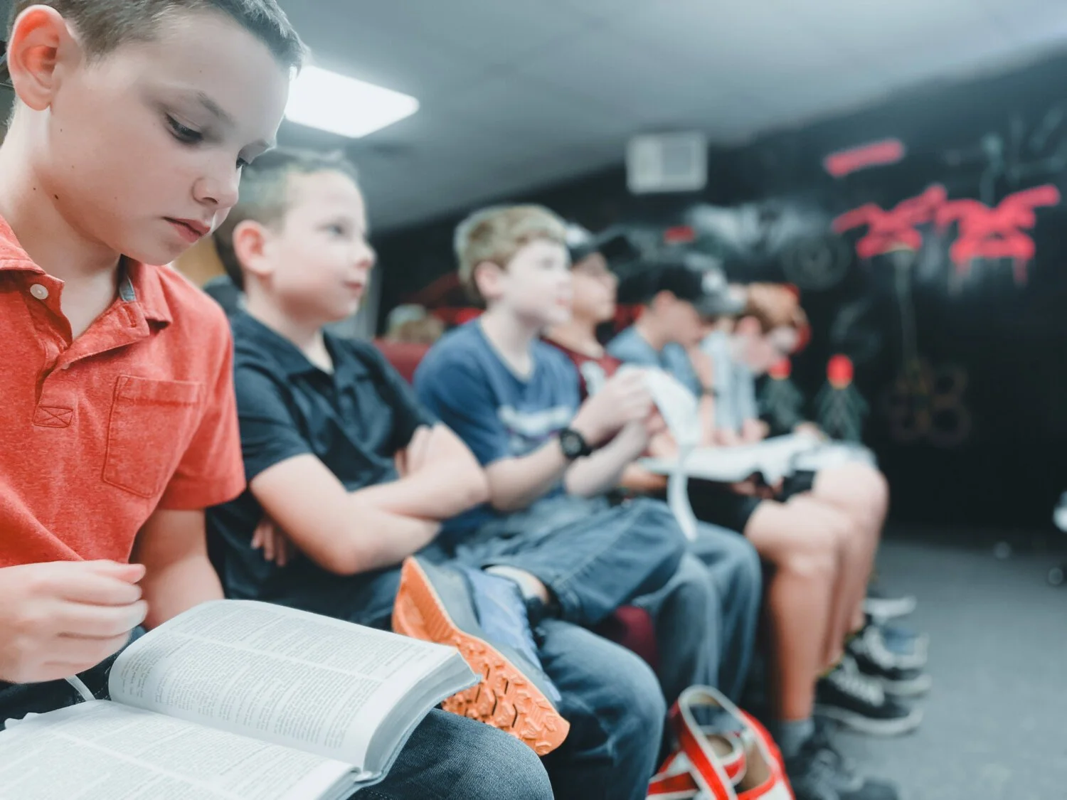 Young boys seated in a row, reading books or looking at their phones in a classroom or club setting with a black wall with red graffiti in the background.