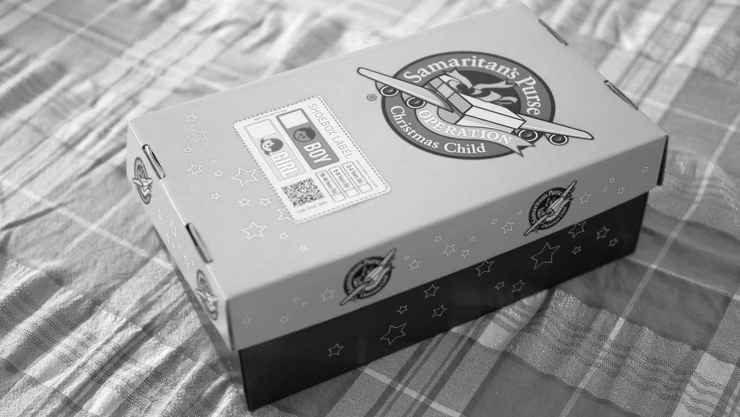A closed shoe box with a Christmas design on it, featuring a plane with a banner reading "Samaritan's Purse Christmas Child." The box has star patterns and a label indicating it is for a boy or girl, with scanning and handling instructions, and is placed on a plaid fabric surface.