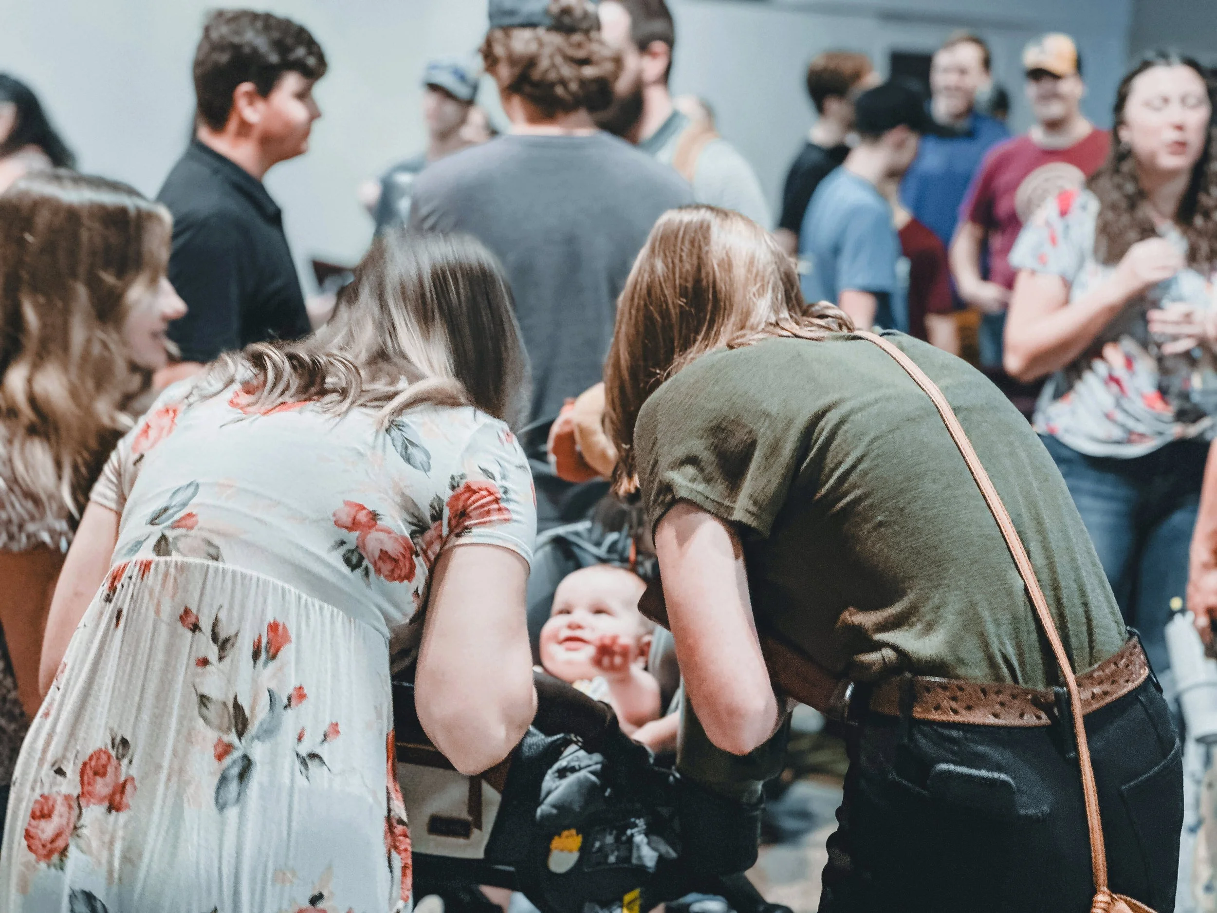 A crowd of people, including a woman in a floral dress, are gathered around a table in what appears to be an indoor event. The focus is on the group engaging with a baby seated at the table, with other attendees standing and talking in the background.