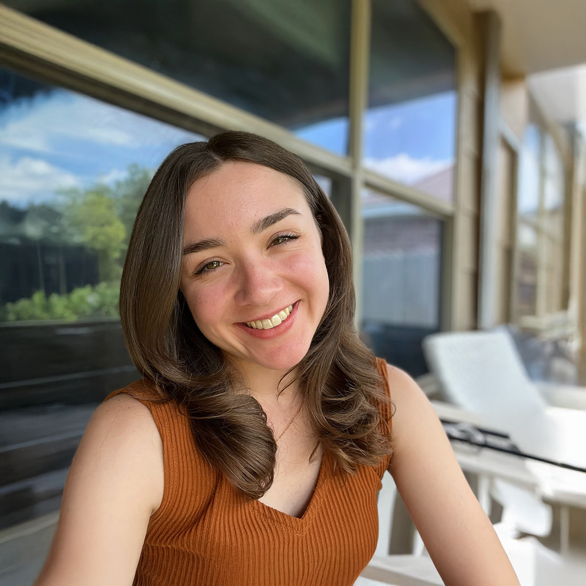 A young woman with shoulder-length brown hair smiling outdoors on a sunny day, wearing a sleeveless orange top, sitting in front of a window showing greenery and a blue sky.