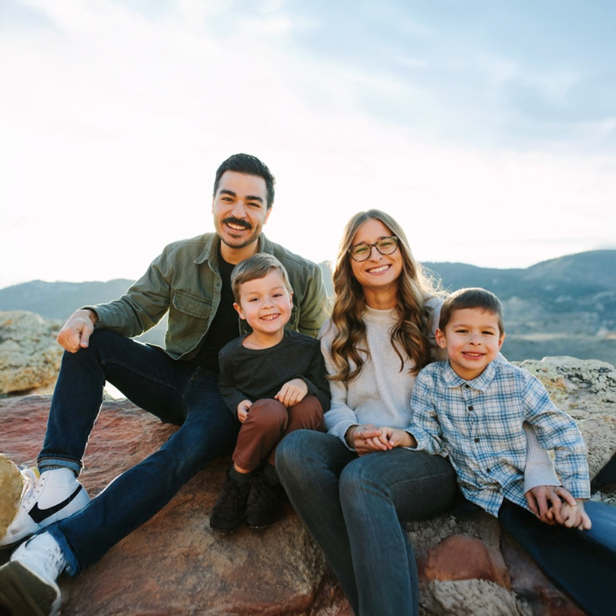 A smiling family of four sitting outdoors on rocks with a mountain landscape in the background, during daytime.
