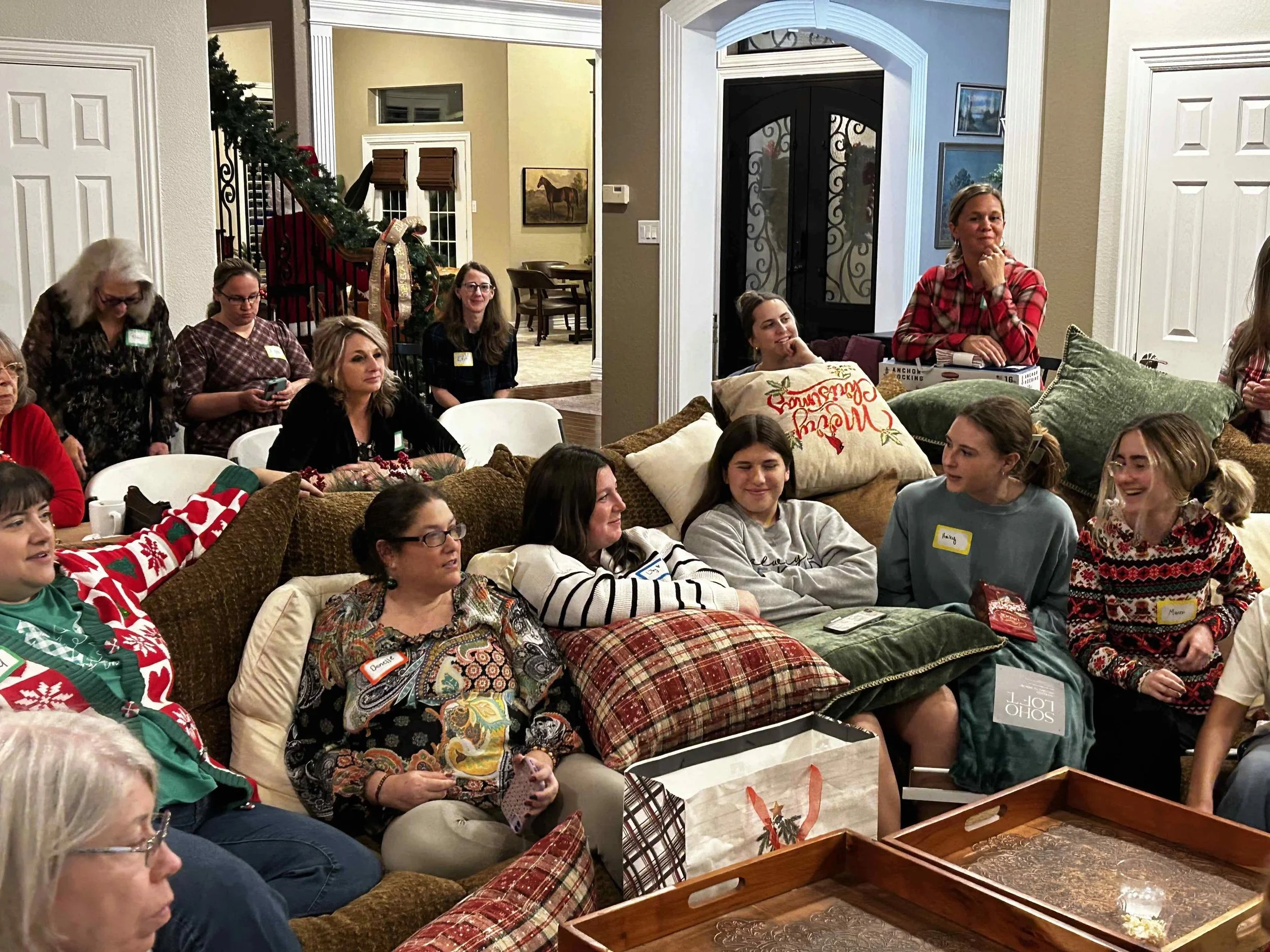 Group of women gathered in a living room decorated for Christmas, some sitting on a couch with holiday pillows, others standing or sitting around, with Christmas gifts and decorations visible.