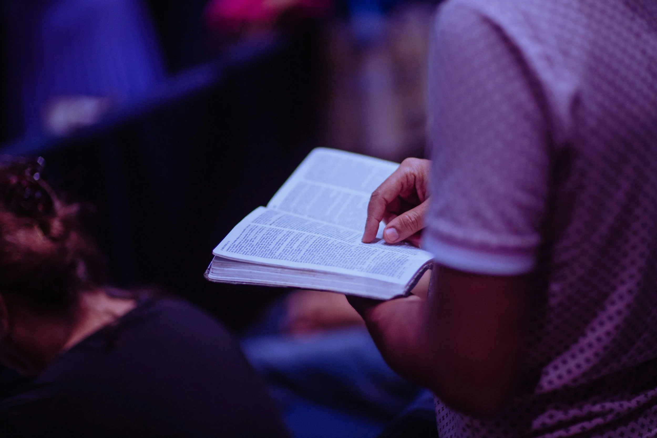 Person holding an open Bible or book, with other people around, in a dimly lit setting.