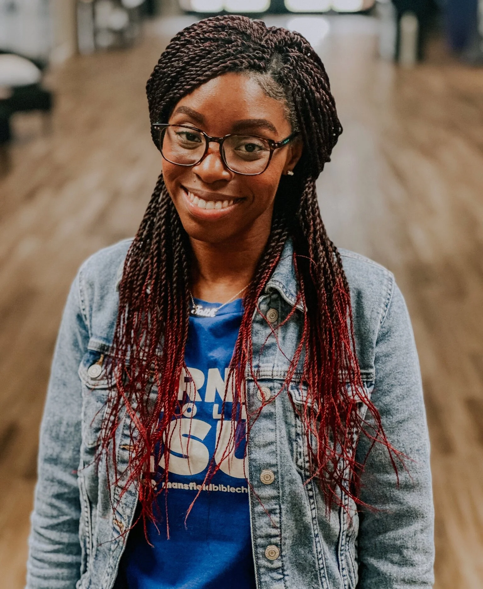 A smiling woman with glasses and long braided hair standing indoors, wearing a denim jacket and a blue T-shirt with white and red text.