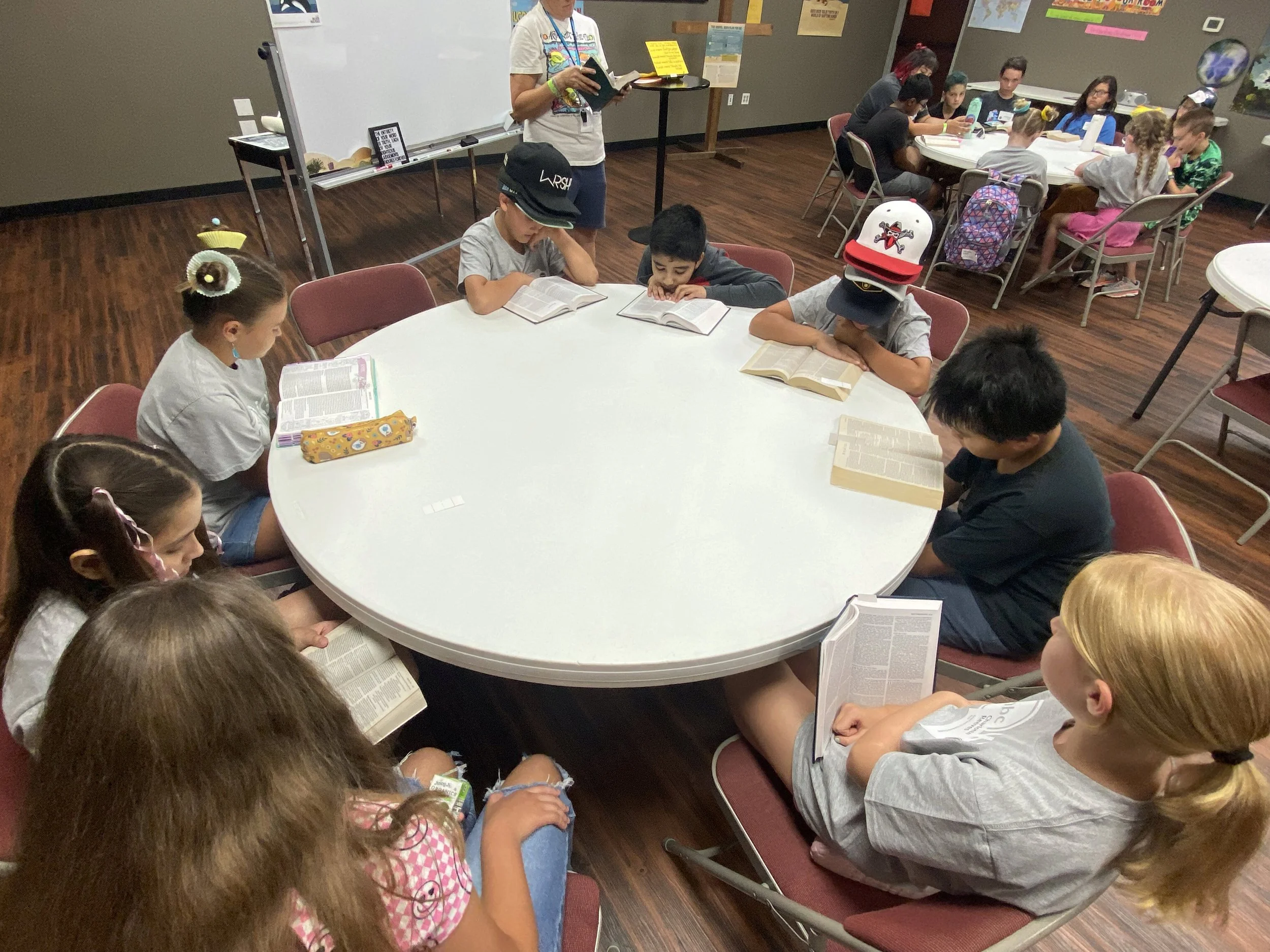 Children sitting at a round table reading from books while a woman stands in the background reading from a book to the group. Additional children are seated at a table in the background.