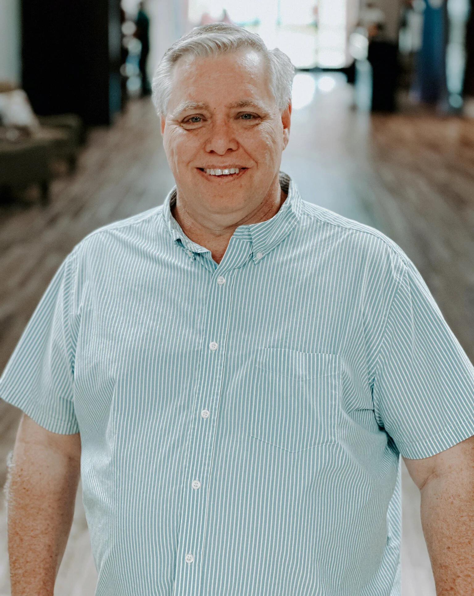 Close-up of an older man smiling, wearing a light blue, striped, button-up shirt, standing in a well-lit room with blurred background.