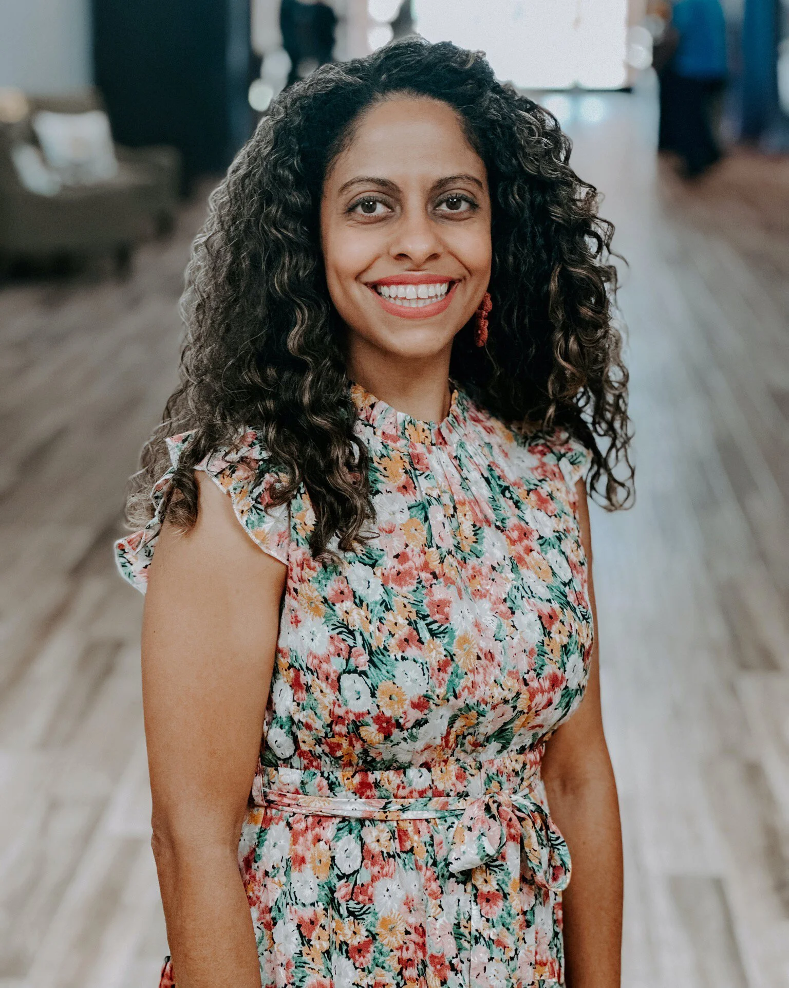 A woman with dark, curly hair smiling at the camera, wearing a colorful floral dress. She is in an indoor setting with blurred background elements.
