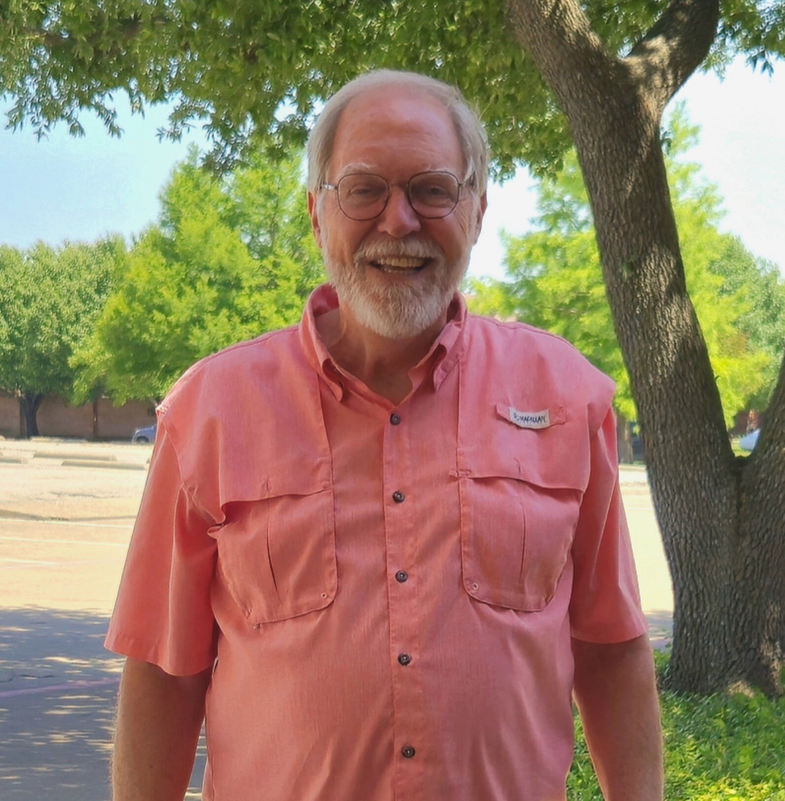 Smiling older man with glasses and a beard, wearing a pink button-up shirt, standing outdoors with trees and a parking lot in the background.