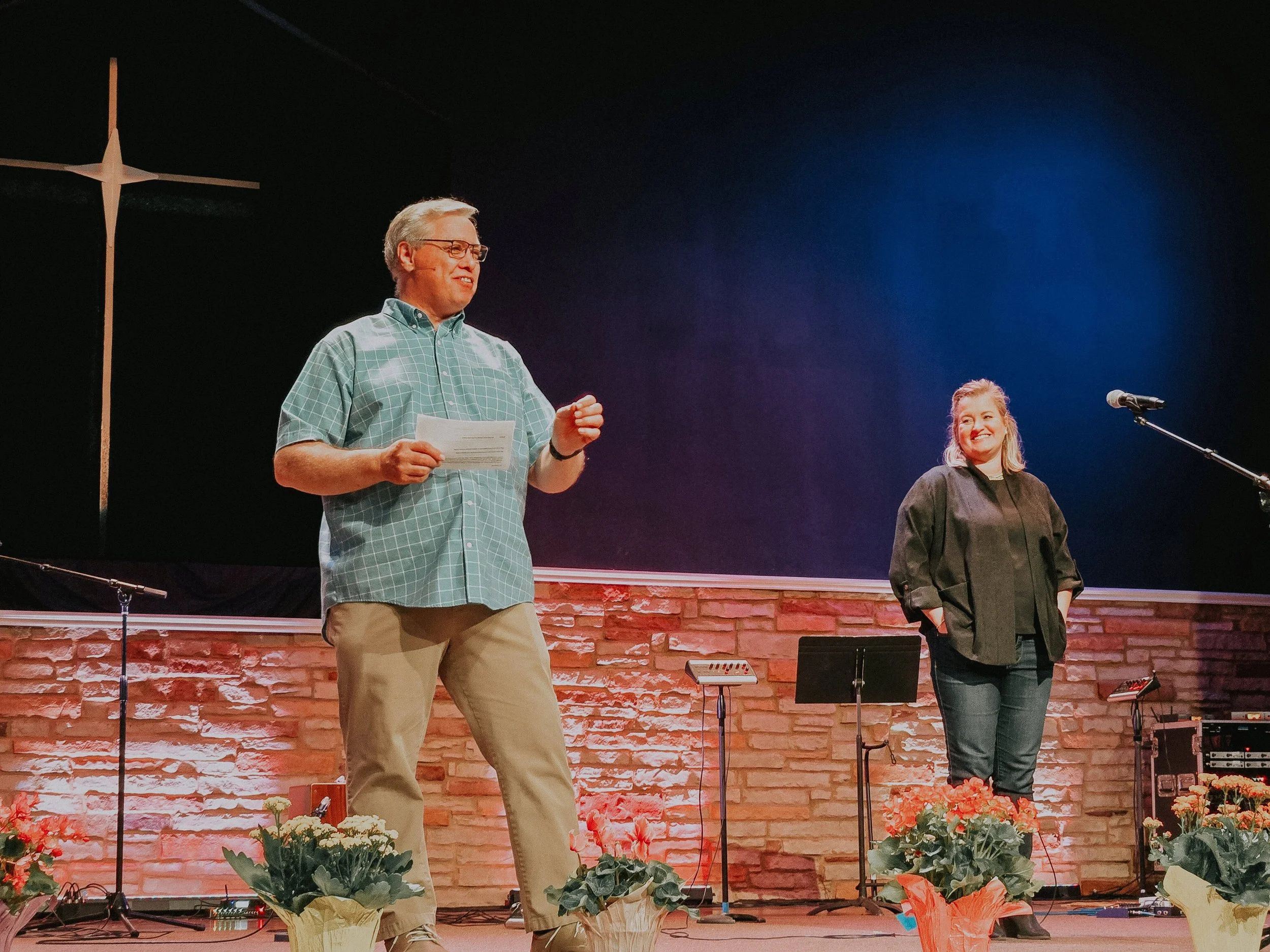 A man wearing glasses in a checkered shirt speaking into a microphone, with a woman in a black outfit standing nearby on a stage with a brick wall background and flowers at the front.