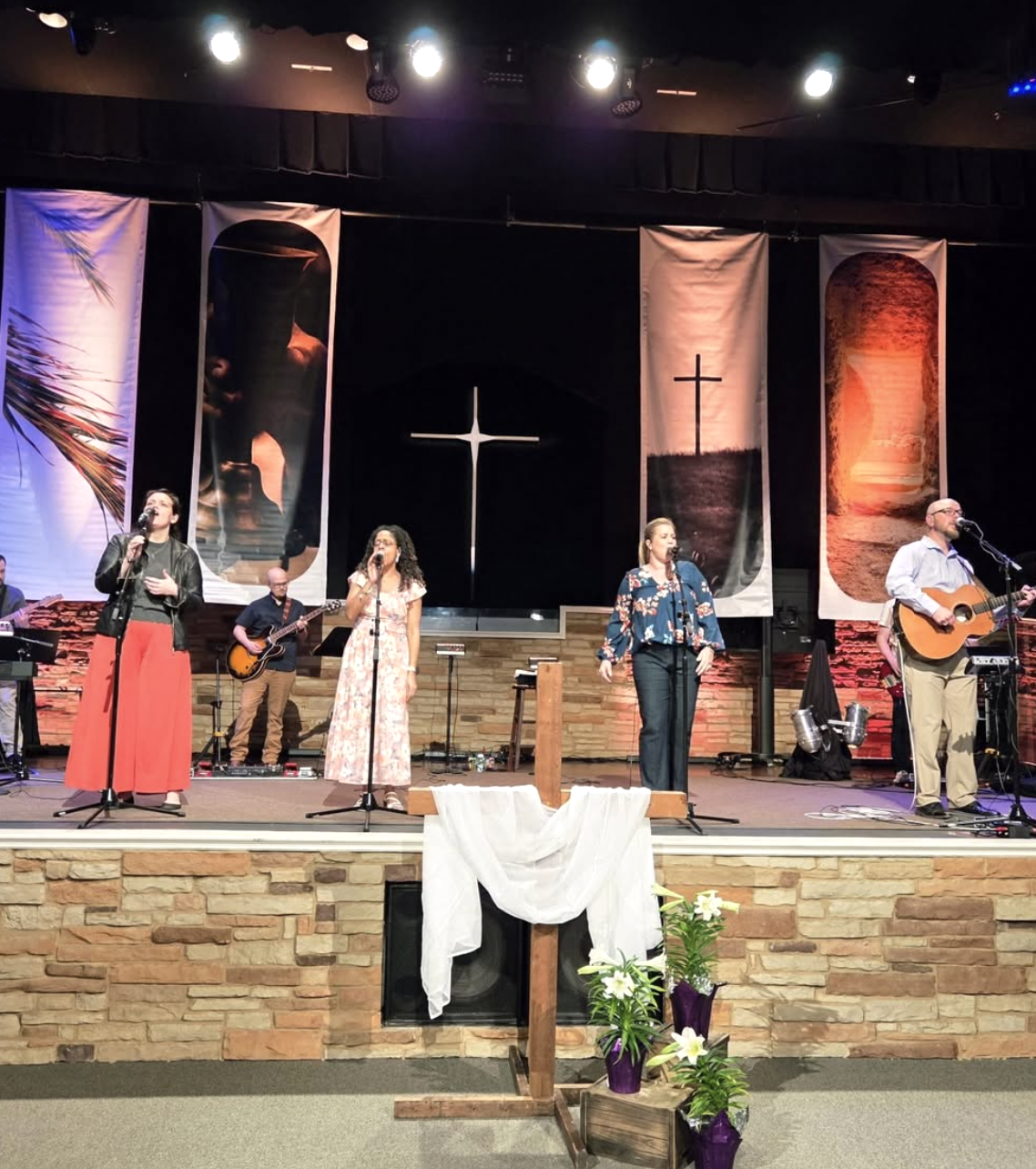 A church stage with five musicians performing, three women and two men, with a cross in the background and religious banners, and flowers in front.