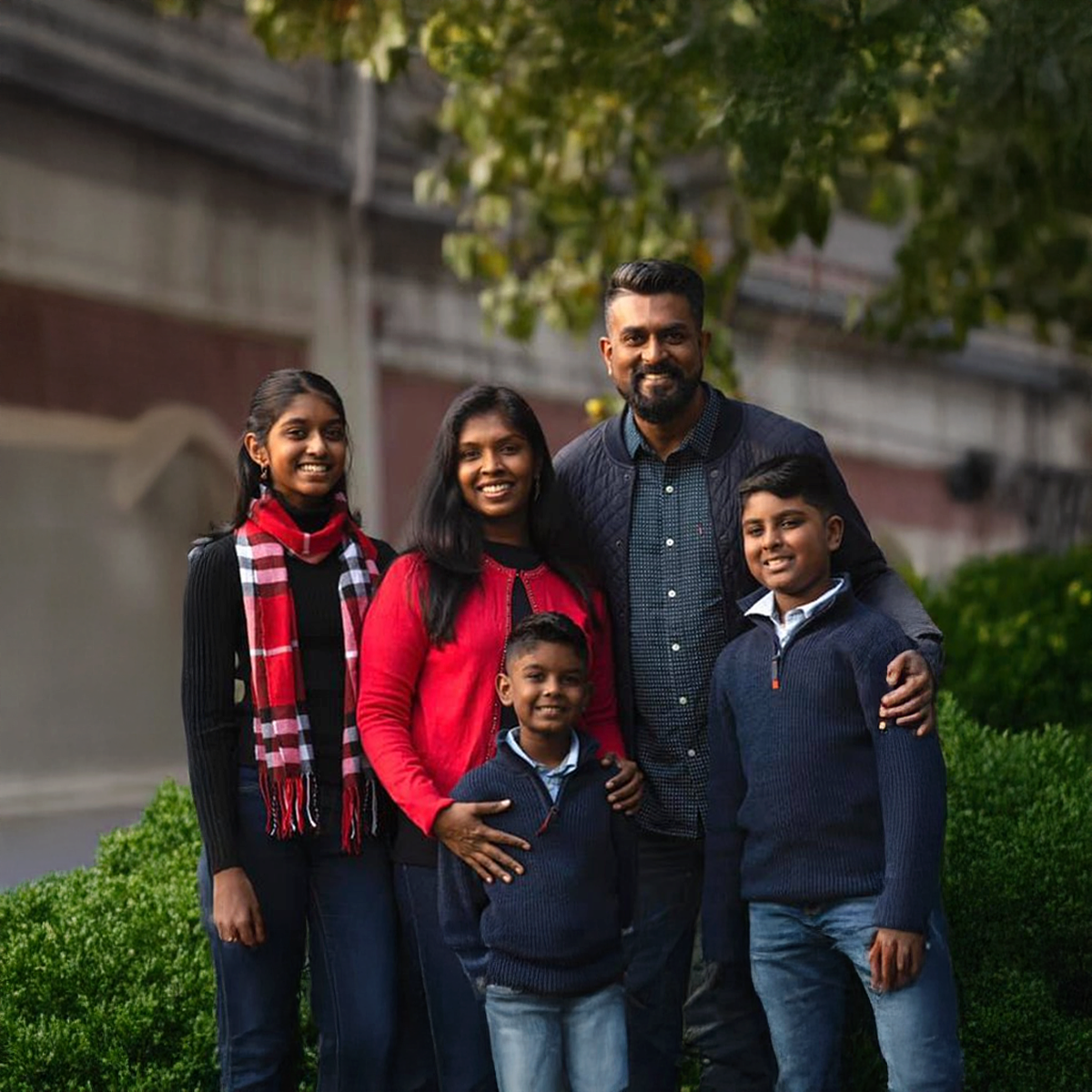 Family of five posing outdoors, with a man and woman surrounded by three children, all smiling at the camera, with greenery and trees in the background.