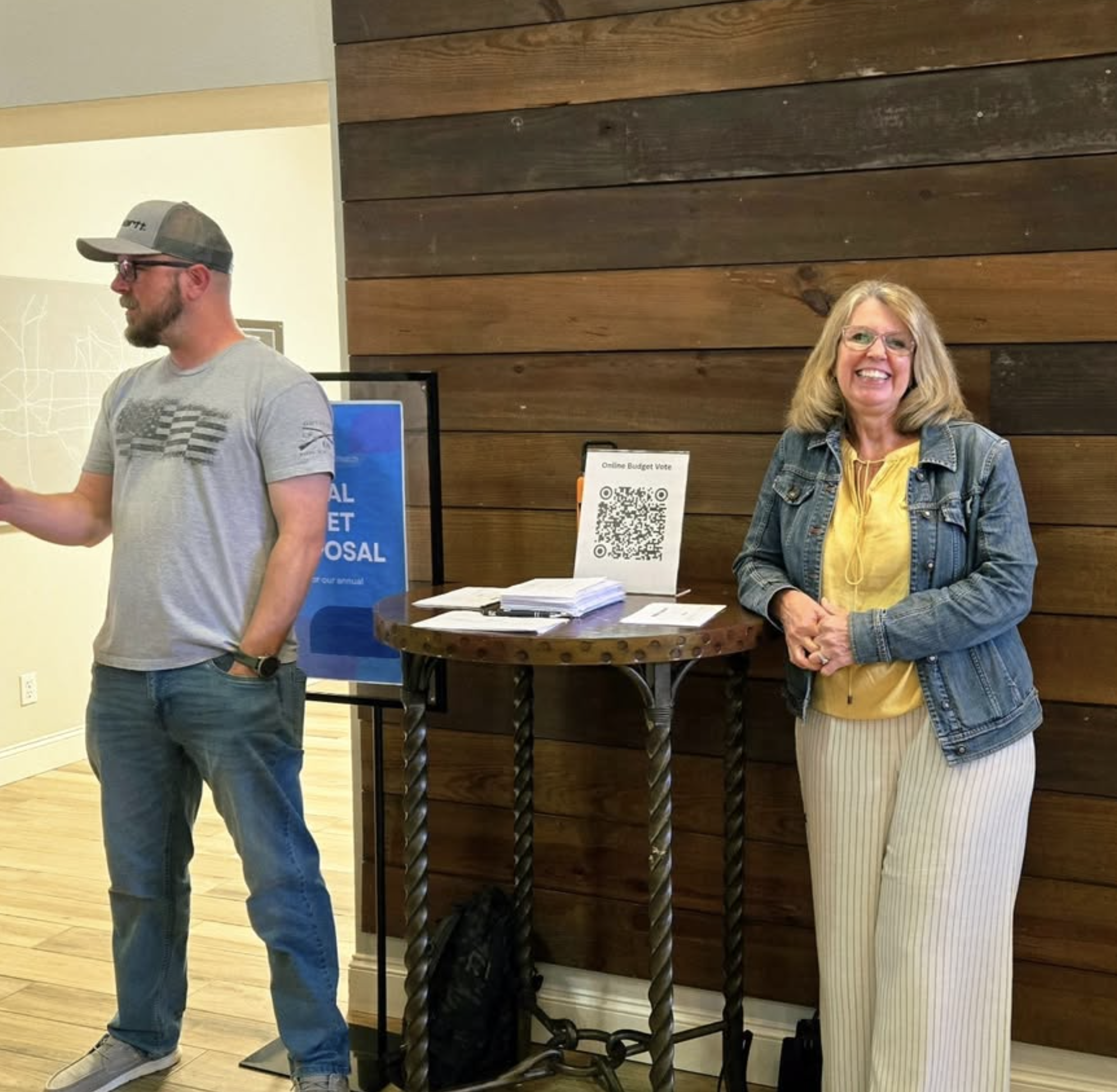 A man wearing a cap, glasses, a gray T-shirt, and jeans is standing with his head turned to the side. A woman with glasses, blonde hair, and a big smile is standing and facing forward. The background features wooden paneling on the wall and a table with papers and a QR code sign.