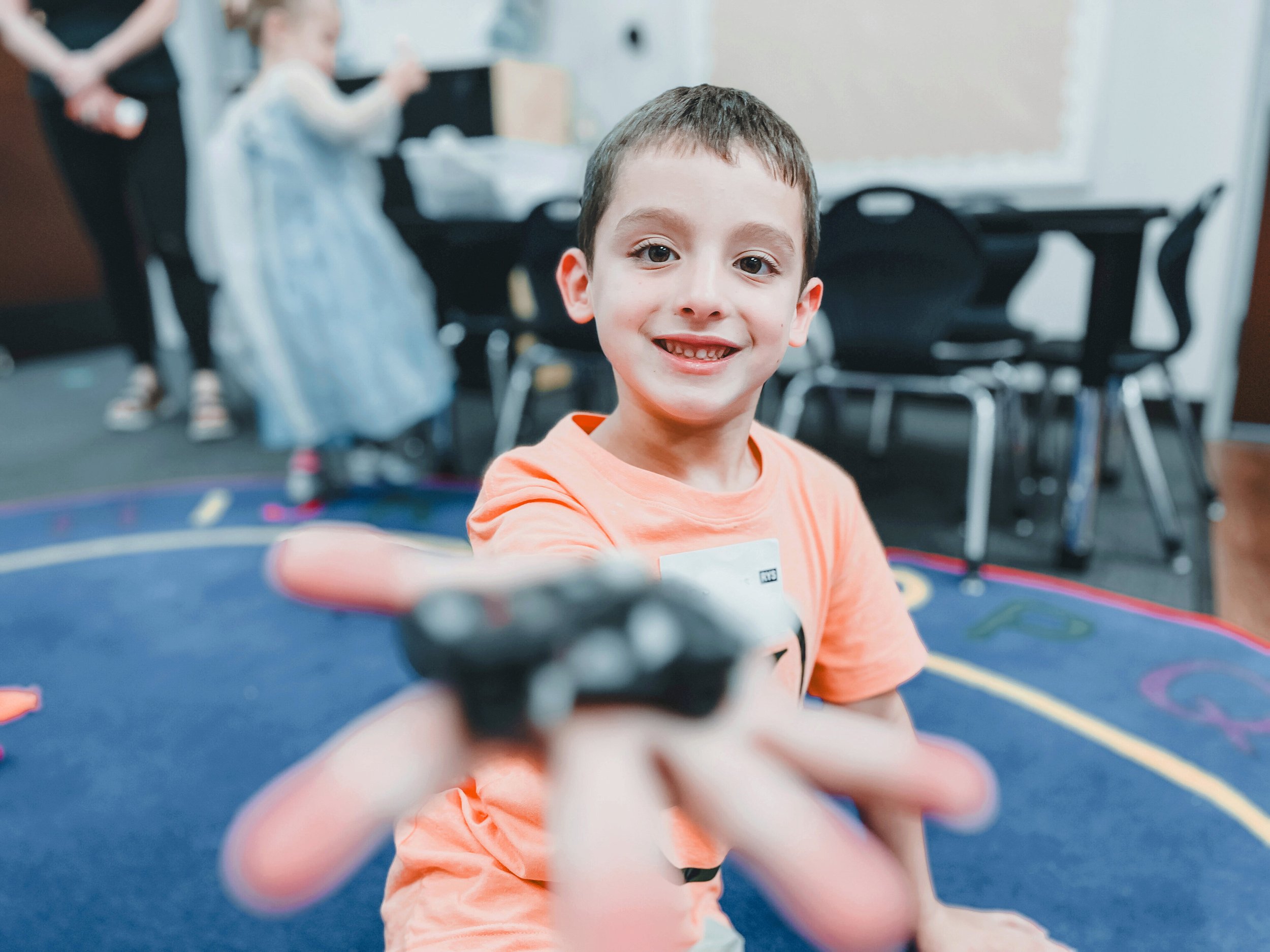 Smiling young boy reaching towards the camera with a blurry background of other children and furniture in a classroom or play area.