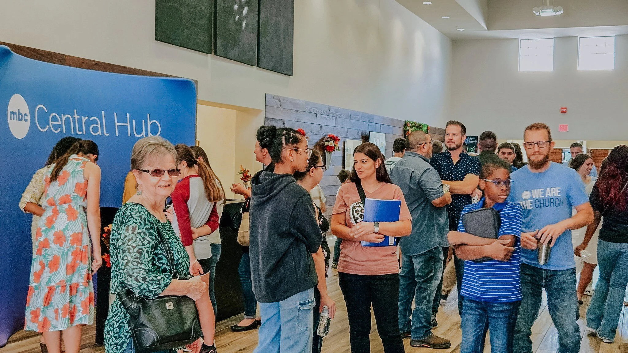 People standing in line at an indoor event, with a large blue sign reading 'mbc Central Hub' on the left side. The background shows a wall with framed pictures and decorative flower arrangements.
