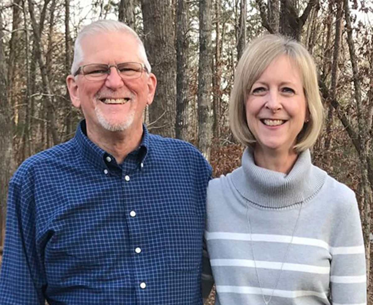 An older man and woman standing outdoors in front of trees, smiling.