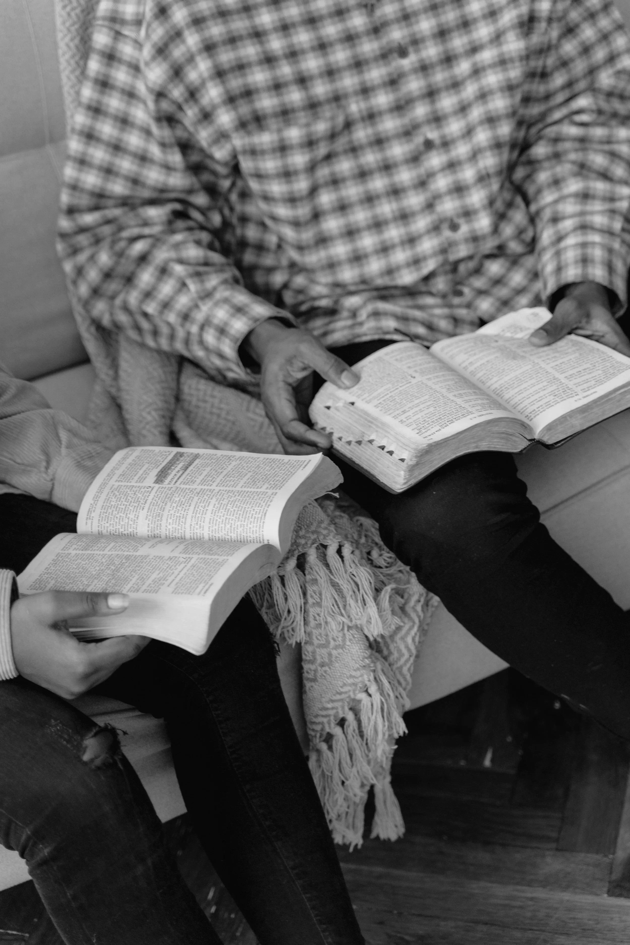 People sitting on a sofa reading opened books, one person wearing a checkered shirt and jeans, with a textured blanket on their lap.