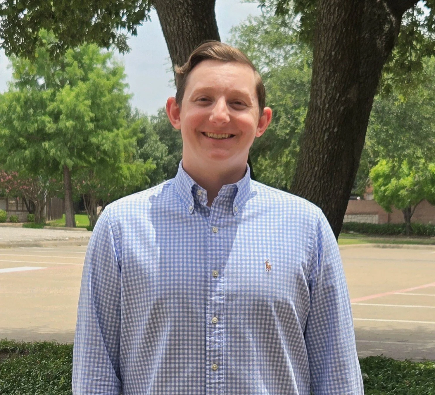 A young man with short brown hair smiling, wearing a blue and white checkered button-up shirt with a small logo of a horse on the chest, standing outdoors near a large tree with greens and houses in the background.