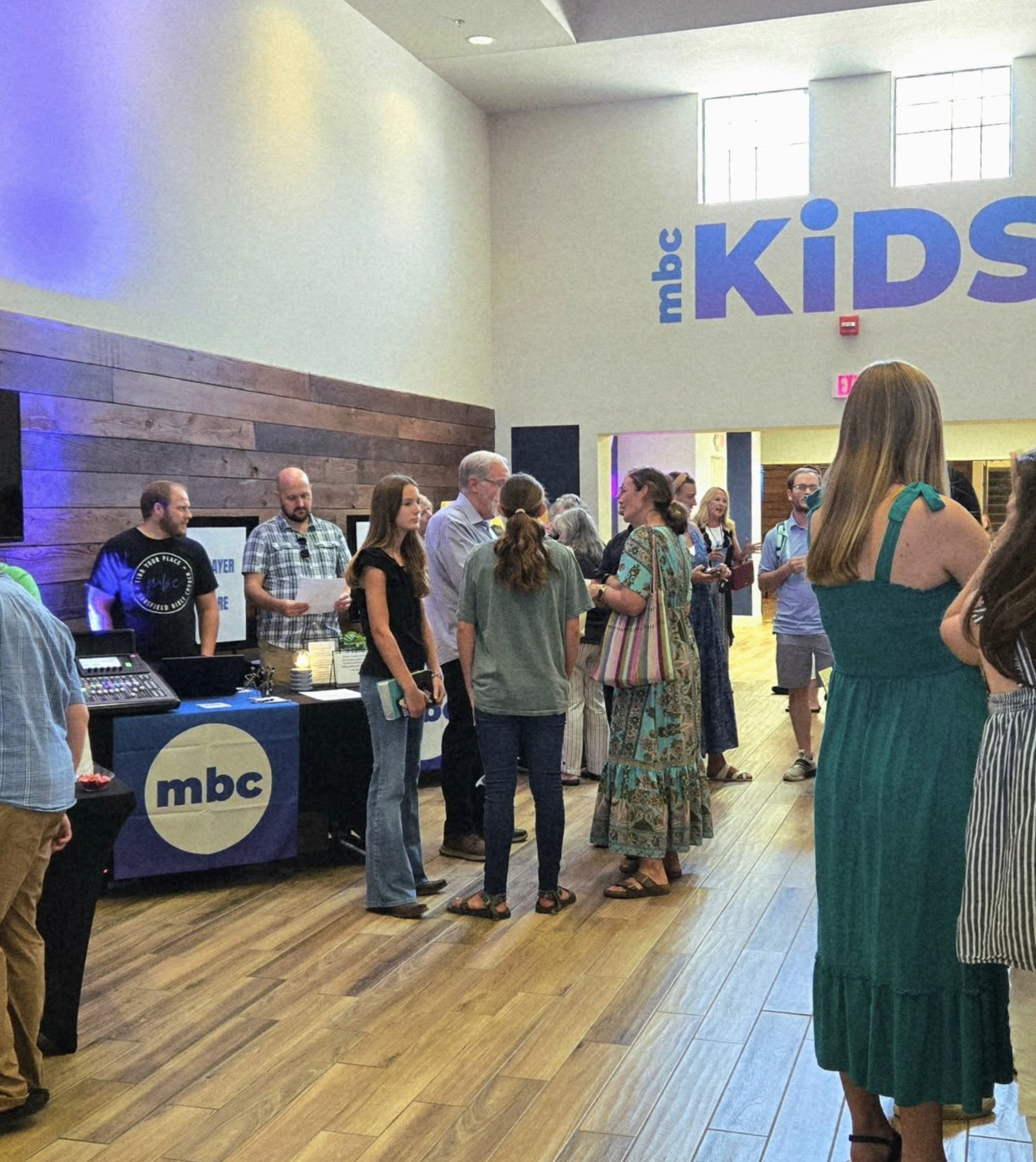 People standing and talking inside a building with a high ceiling, light-colored walls, and wooden floors. A table with a blue cloth displaying the 'mbc' logo is visible. Large windows let in natural light. The scene appears to be a gathering or registration area.