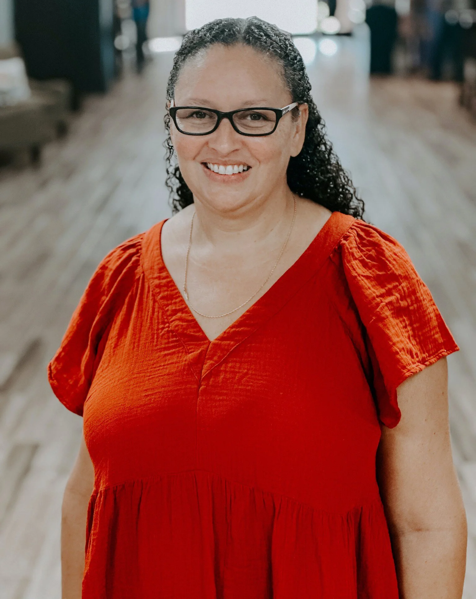 A woman with glasses and curly hair, wearing an orange dress, smiling in an indoor space with blurred background.