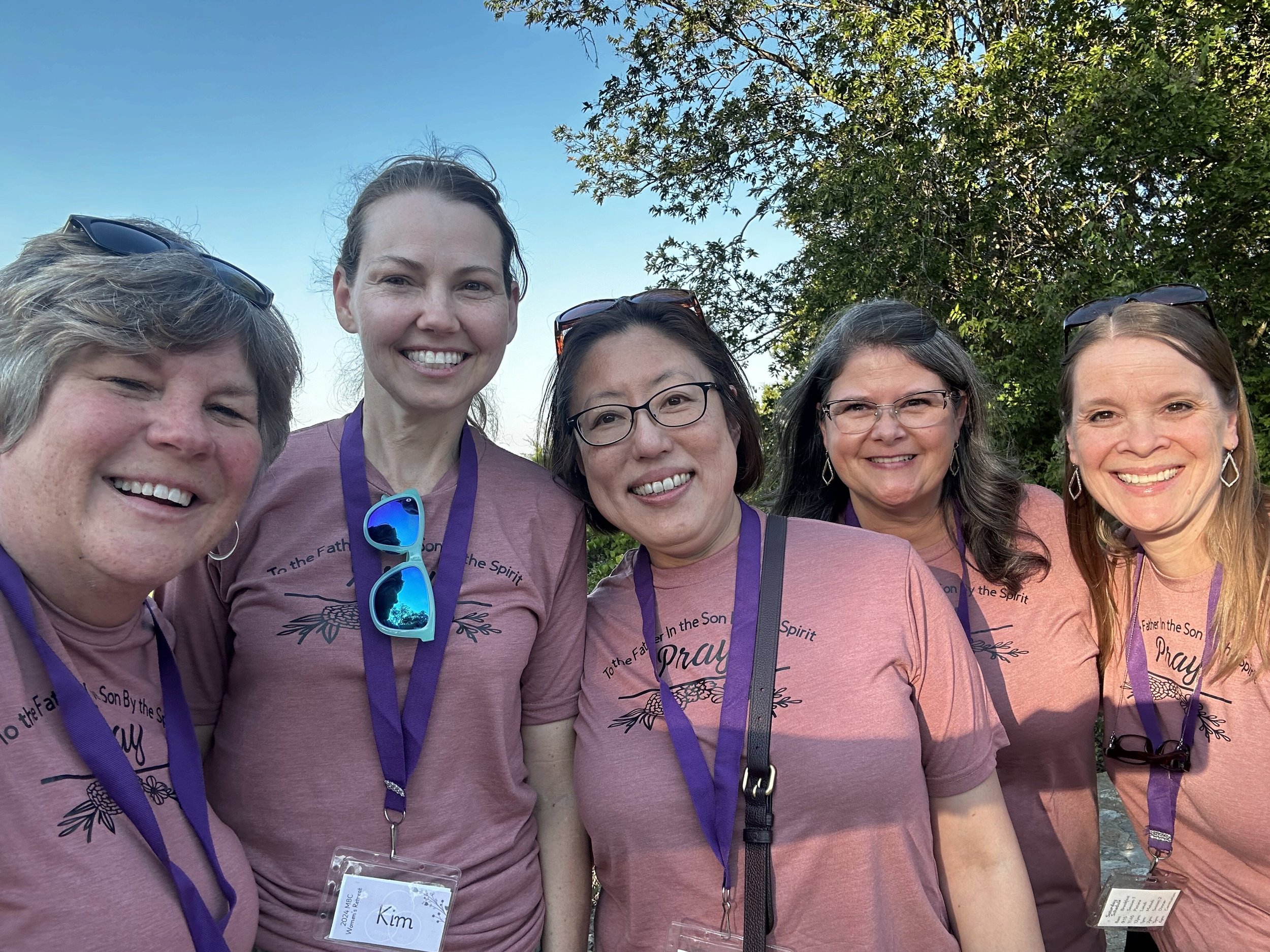 A group of five women smiling outdoors, wearing matching pink T-shirts and purple lanyards with name tags, standing under a tree on a sunny day.