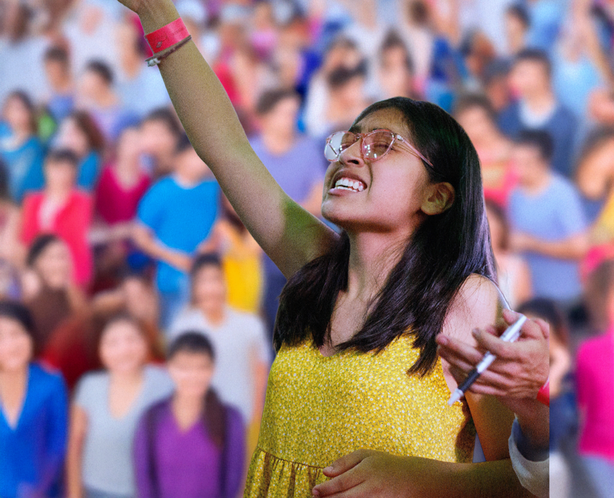 A young girl with glasses and a yellow dress appears emotional with eyes closed and hand raised, in a crowd of people, possibly at a public event or rally.