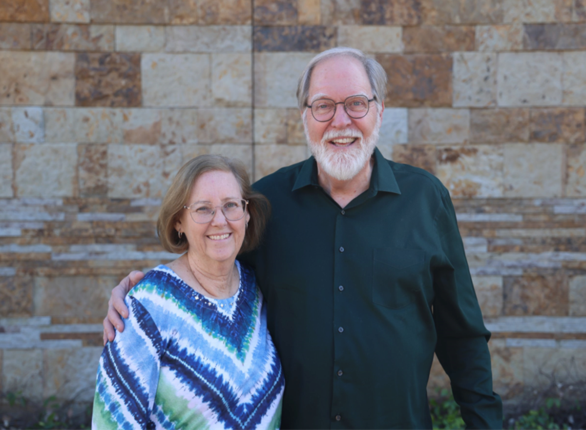 An elderly couple standing outdoors with a stone wall background, smiling, the woman wearing glasses and a colorful patterned top, the man with glasses and a black shirt.