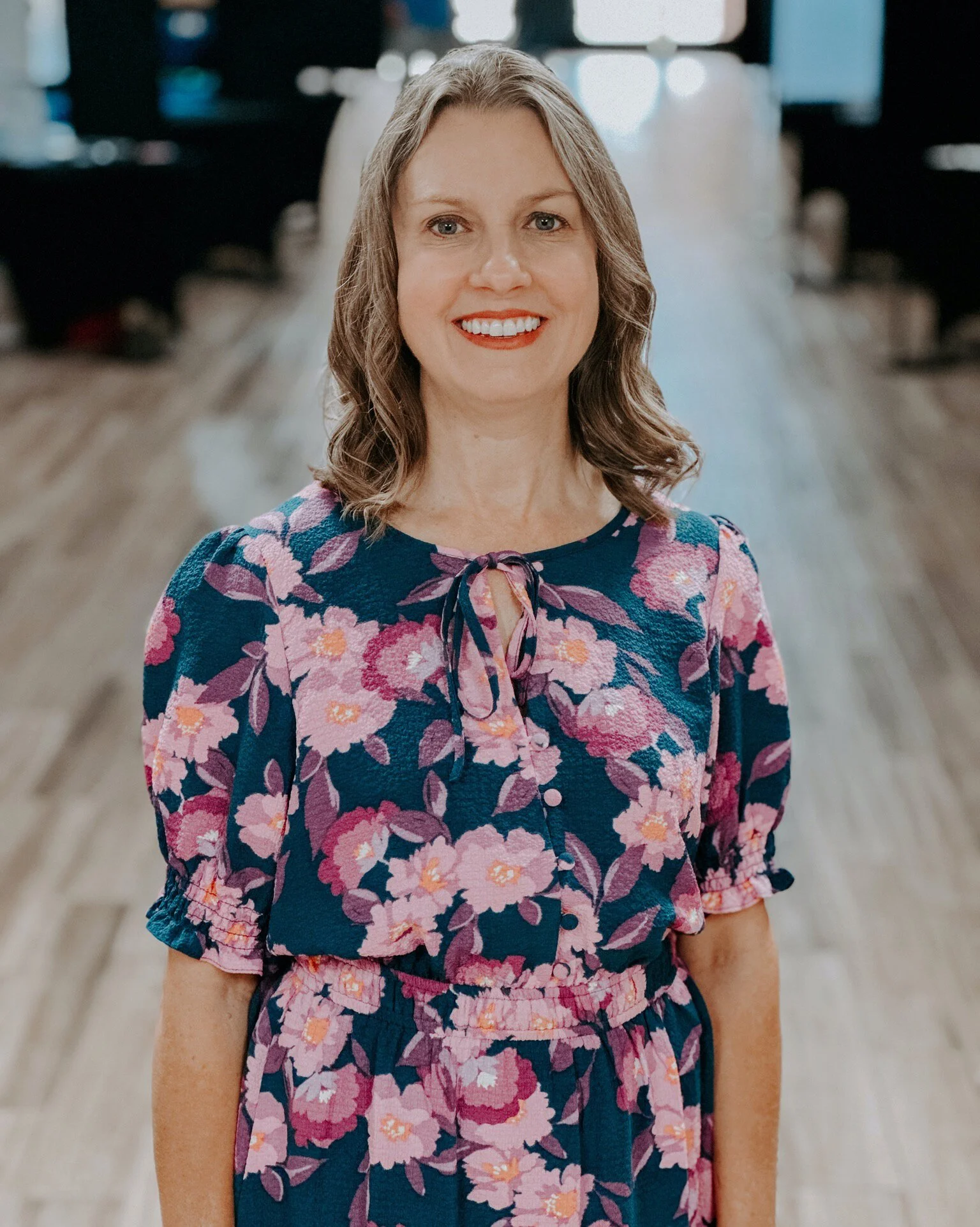 A woman with shoulder-length gray hair and blue eyes smiling, wearing a floral dress with pink, purple, and blue flowers, on a wooden floor in a room with dark furniture and a window in the background.