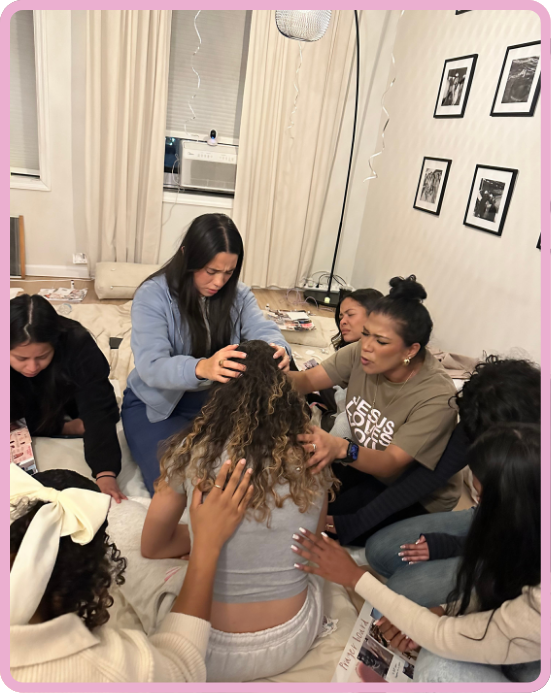 Group of women sitting on the floor in a circle, engaging in a prayer, one woman in the center has her head bowed and hands raised, others touching her shoulders or head.