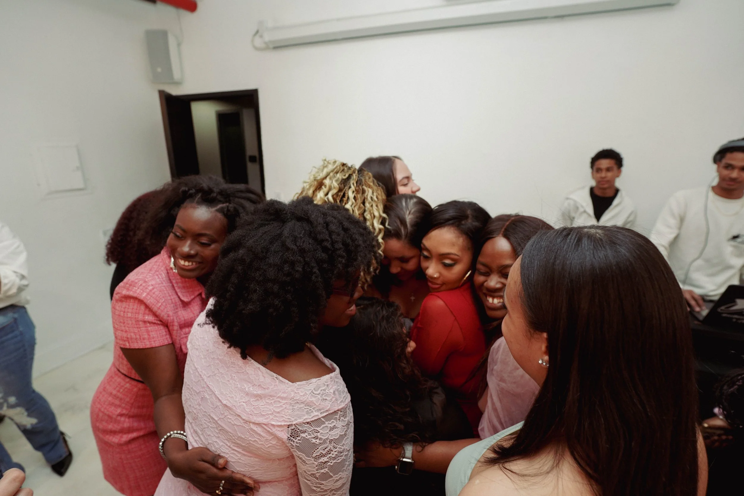 A group of women with diverse backgrounds is gathered together, smiling and embracing in an indoor setting. In the background, some men are standing and observing. The women appear to be happy and close, sharing a moment of connection.