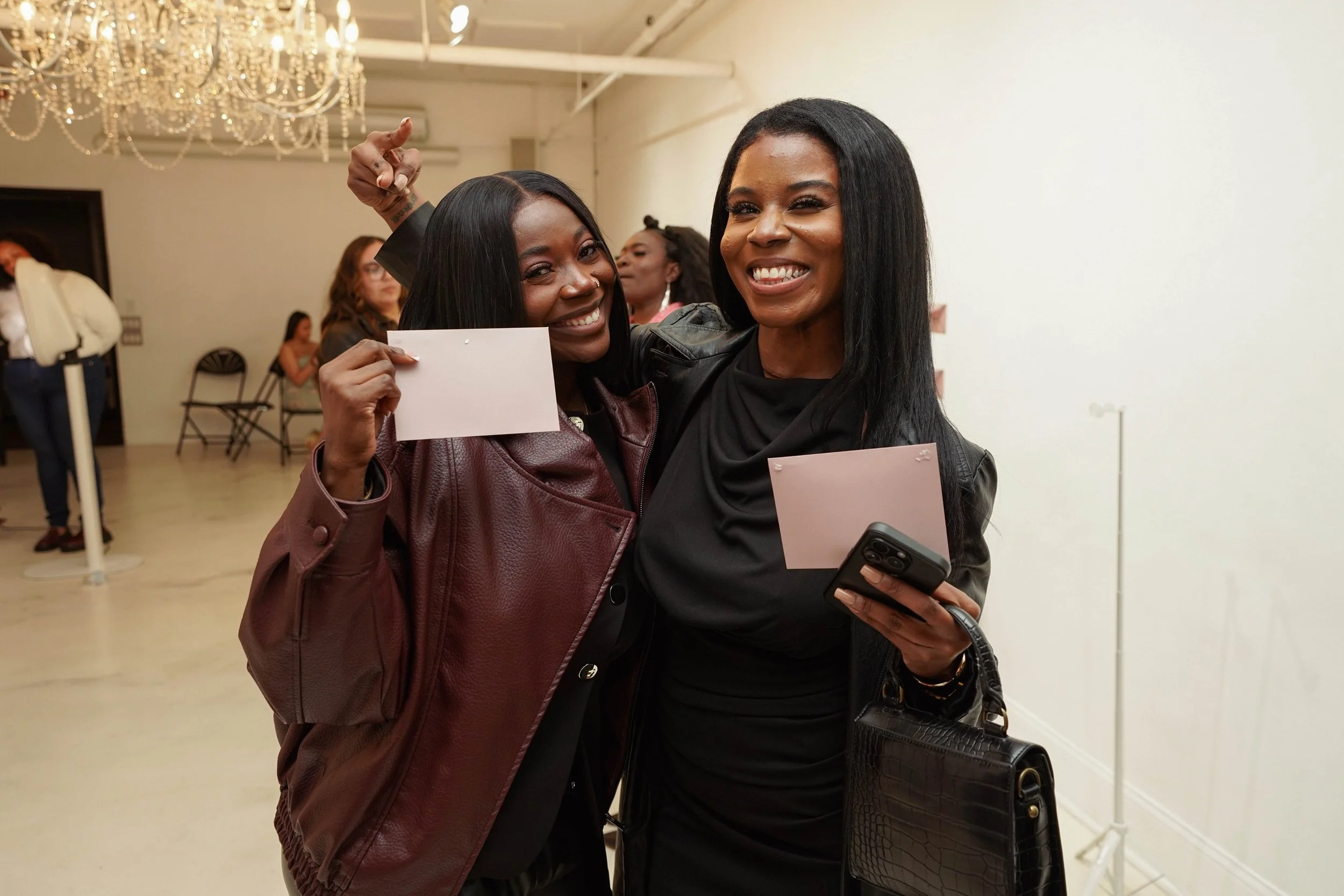 Two women smiling, embracing, and holding envelope and phone at an indoor event, with other people and a chandelier in the background.