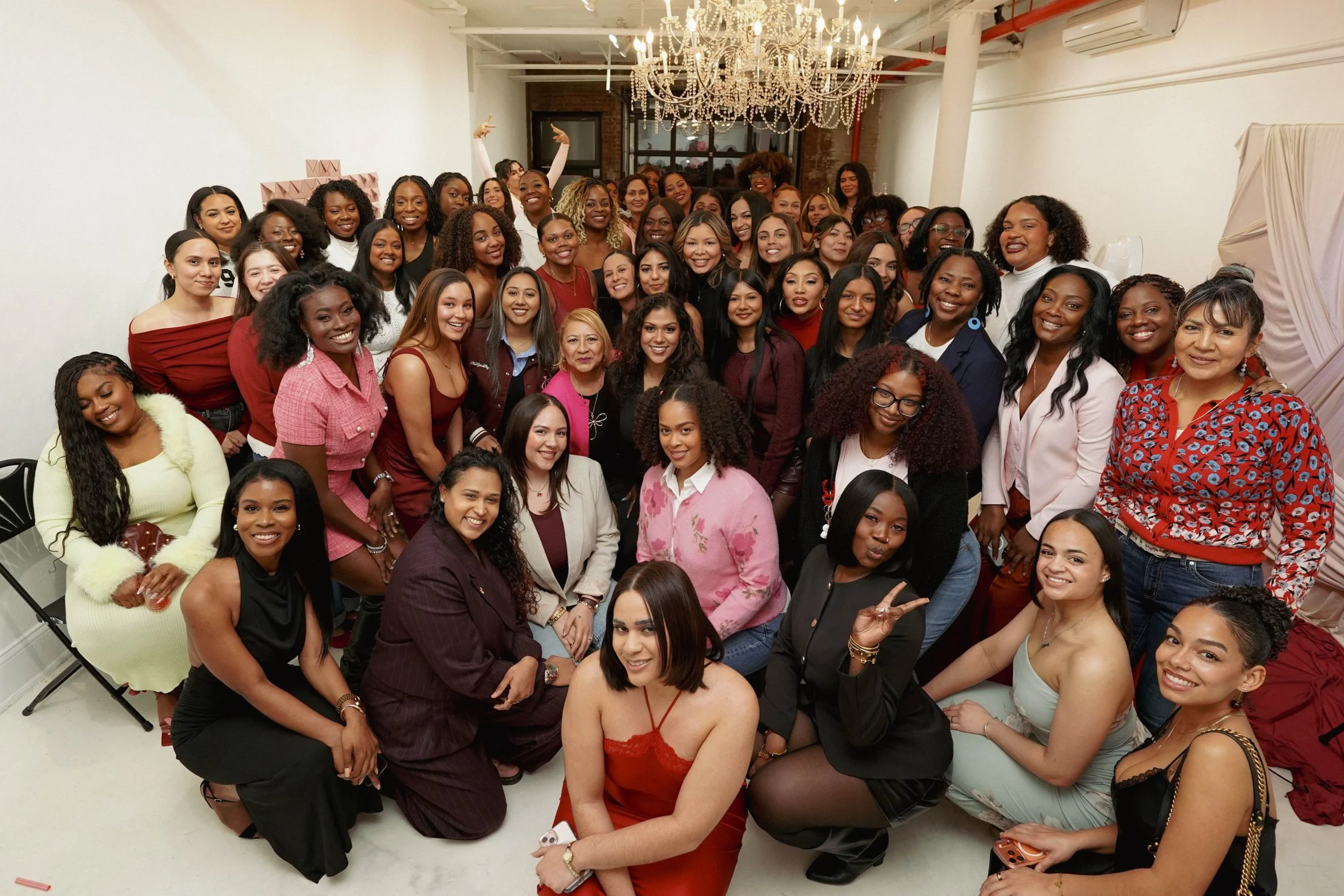 Group of women gathering indoors for a celebration or event, smiling and posing for the photo, with some seated and some standing, in a room decorated with a chandelier.
